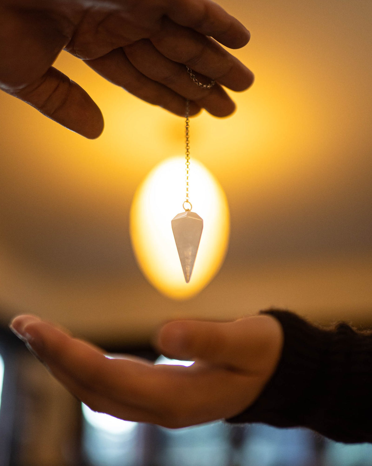 A clear quartz pendulum hangs from a fine chain, lit from behind by warm light, above an open hand.