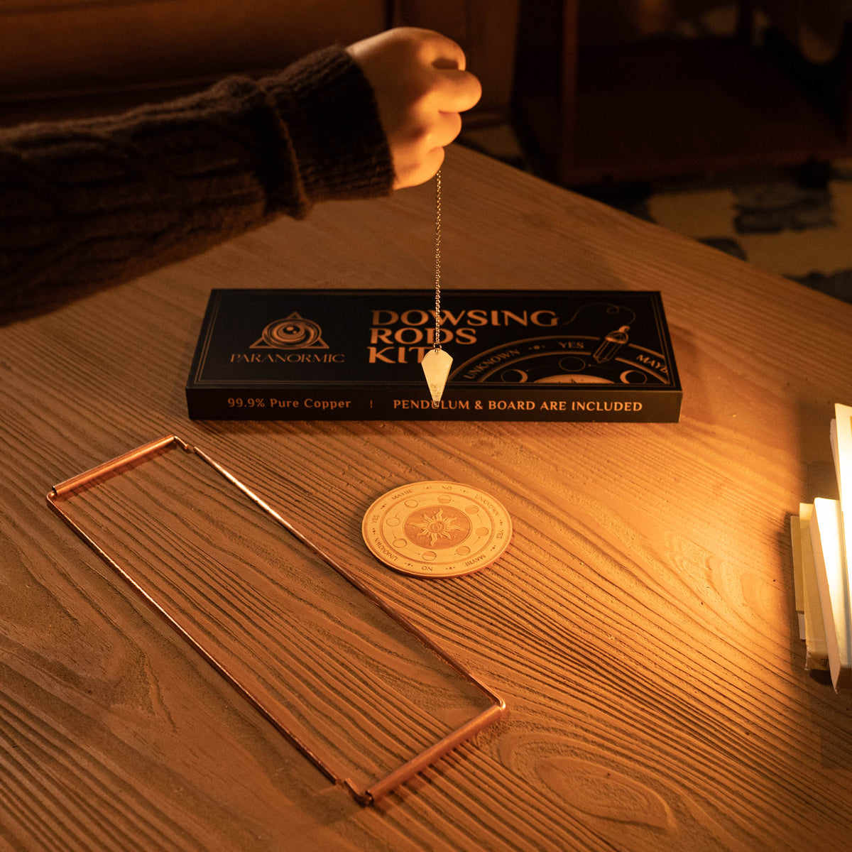 Real Paranormic copper rods and wooden answer board on a table, with a quartz pendulum held beside the printed gift box.