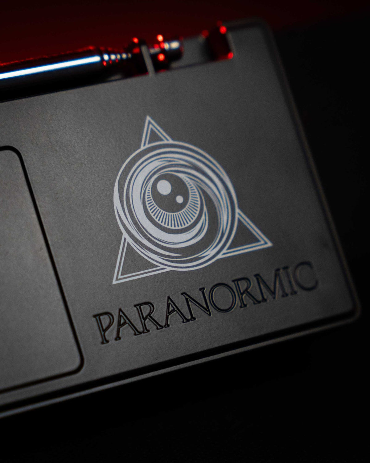 The embossed Paranormic eye-and-triangle logo on the back of the Spirit Box, lit in low red light.